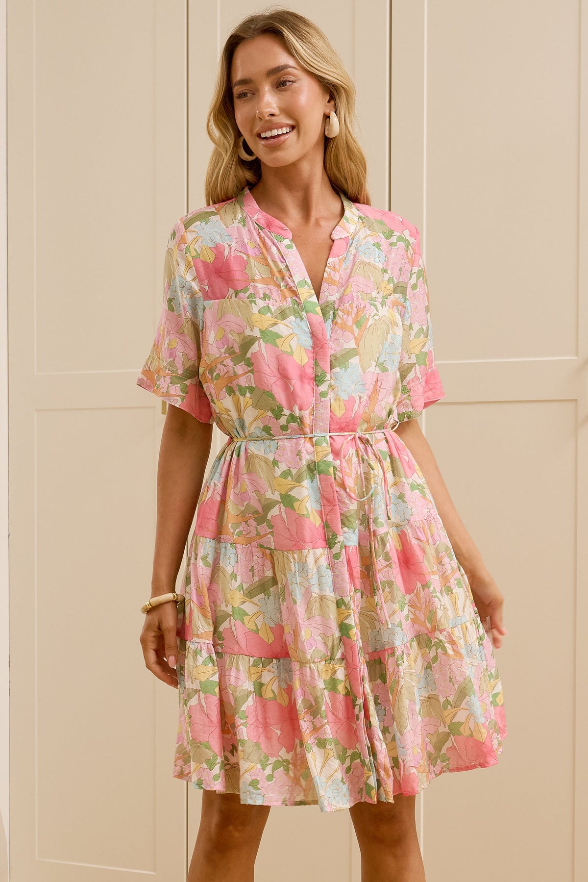 A woman modeling a pink and green floral short-sleeve dress with a tie waist, standing in a brightly lit room with cream-colored wooden wardrobes. Her reflection is visible in a mirror on the left.