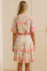 A woman stands with her back to the camera wearing a short-sleeved floral dress with a pink waist tie, standing in a room with cream-colored built-in wardrobes and a large floor mirror showing her reflection.