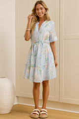 A woman stands in a bright room wearing a light blue floral mini dress with short sleeves and a tie waist, paired with white slide sandals. A large mirror on the left reflects her side profile, and cream-colored wardrobe doors and a white vase are visible in the background.