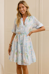 A woman stands in a bright room with cream-colored cabinets and a wooden floor, wearing a light blue and white floral print mini dress with short flutter sleeves and a tie waist. She is standing next to a large white vase, with her reflection visible in a mirror on the left.