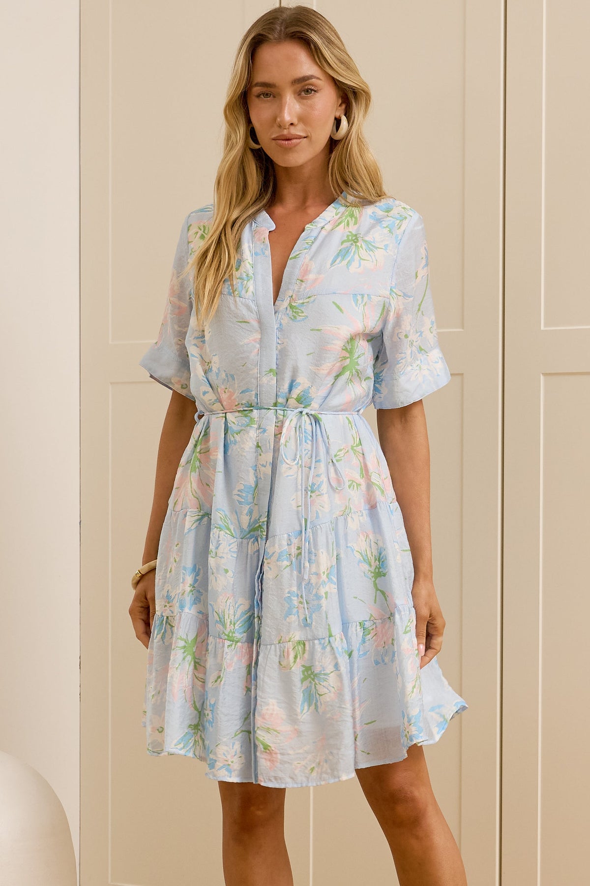 A woman stands in a bright room wearing a light blue floral short-sleeve dress and beige slide sandals, with a large mirror on the left reflecting her side profile. The background features cream-colored wooden wardrobe doors.