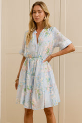 A woman stands in a bright room wearing a light blue floral short-sleeve dress with a tie waist, with a large mirror on the left reflecting her side profile. She is wearing beige slide sandals.