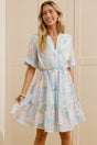 A woman stands in a bright room wearing a light blue floral short-sleeve dress and beige slide sandals, with a large mirror on the left reflecting her side profile. The room has cream-colored paneled walls and doors, light wood flooring, and two large white ceramic vases.