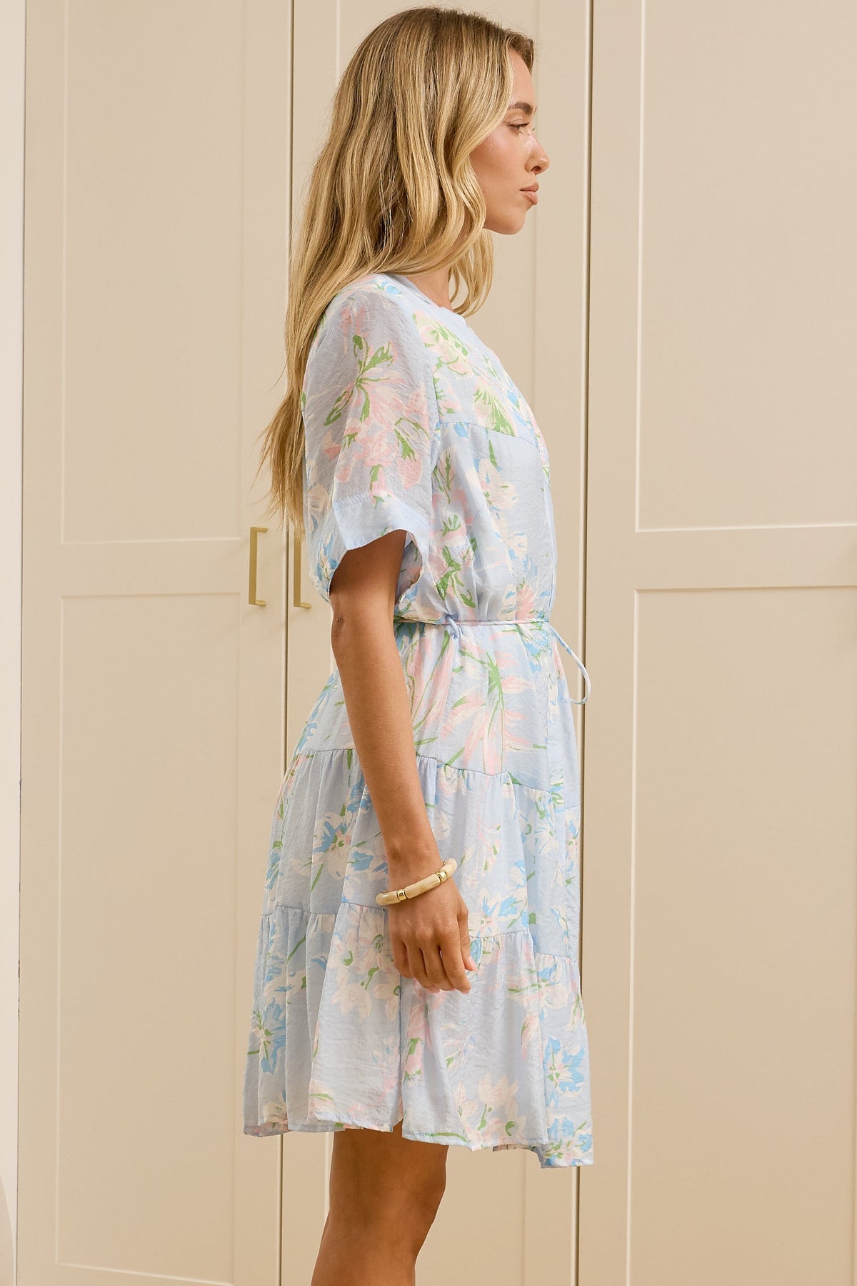 A woman stands in profile wearing a light blue and white floral print short-sleeve dress with a tie waist, standing in a bright room with cream-colored wooden wardrobes and a large mirror showing her reflection.