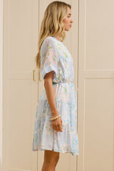 A woman stands in profile wearing a light blue and white floral print short-sleeve dress with a tie waist, standing in a bright room with cream-colored wooden wardrobes and a large mirror showing her reflection.