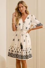 A woman stands in a bright room wearing a cream-colored dress with black geometric patterns and a matching tie belt. She is standing in front of white wardrobe doors and a large mirror that reflects her side profile.