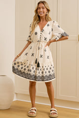 A woman models a cream-colored short-sleeve kaftan dress with a black geometric pattern, standing in a brightly lit room with cream-colored cabinets. A large mirror on the left reflects her side profile.