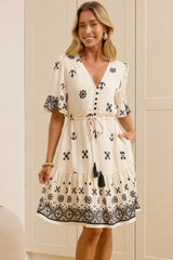 A woman models a cream-colored short-sleeve dress with black bohemian-style patterns, standing in front of a large mirror that reflects her side profile. The room features cream-colored paneled walls, a light wood floor, and two large white ceramic vases.