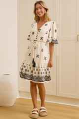 A woman modeling a cream-colored cotton dress with black floral embroidery and a black patterned hem, standing in a bright room with white wardrobes and a wooden floor.