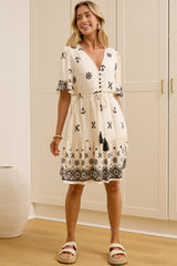 A woman modeling a cream-colored short-sleeve dress with black bohemian-style embroidery and tassel details, standing in a brightly lit room with cream-colored cabinets and a wooden floor.