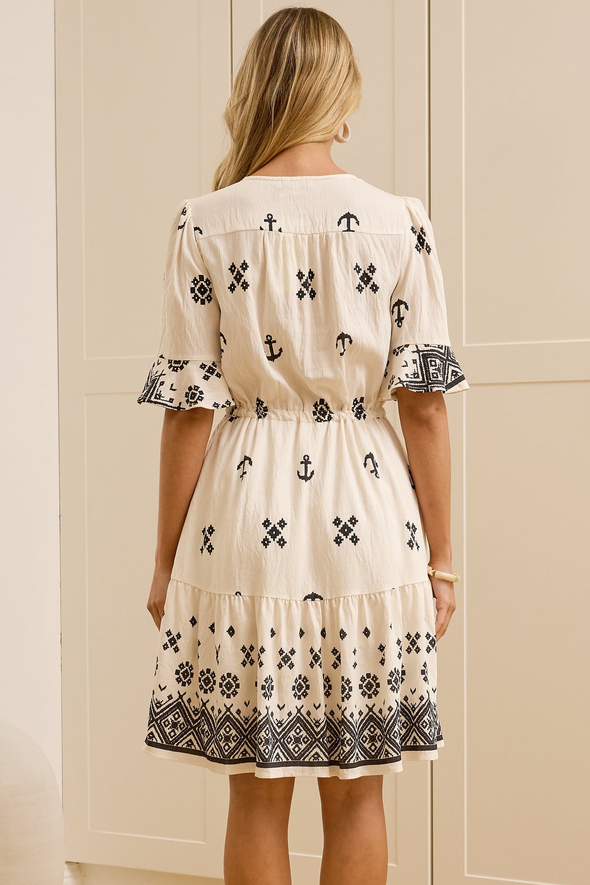 A woman stands with her back to the camera wearing a cream-colored dress with black floral embroidery, in a bright room with cream-colored wardrobes and a large mirror showing her side profile.