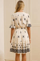 A woman stands with her back to the camera wearing a cream-colored dress with black floral embroidery, in a bright room with cream-colored wardrobes and a large mirror showing her side profile.