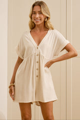A woman modeling a short cream-colored linen romper with a V-neckline and front buttons, standing in front of a cream-colored paneled wall.