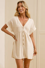 A woman stands in a bright room with light wood floors and white wardrobes, wearing a short cream-colored wrap dress with short sleeves and a front button detail. She is standing in front of a large floor-to-ceiling mirror that reflects her side profile.