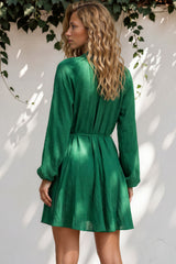 Woman wearing a green dress standing against a white wall with foliage.