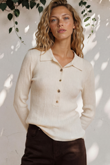 Woman wearing a cream ribbed sweater and dark pants standing against a white wall with greenery.