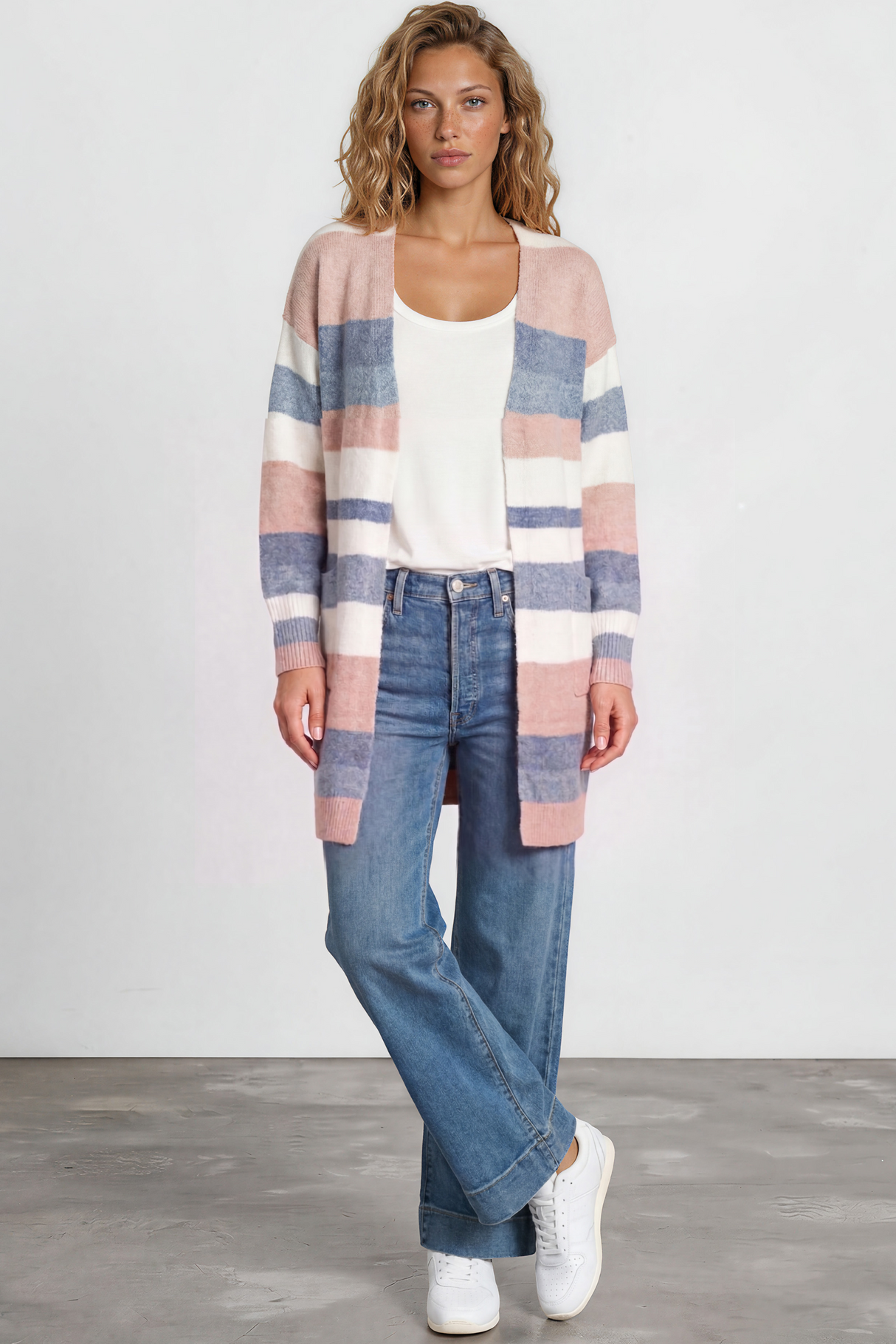 Woman wearing a striped cardigan, white top, blue jeans, and white sneakers on a plain background