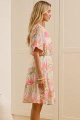 A woman stands in a bright room wearing a short-sleeve tiered floral dress with pink, green, and beige tones. A large mirror on the left reflects her side profile, and cream-colored cabinets line the background wall.
