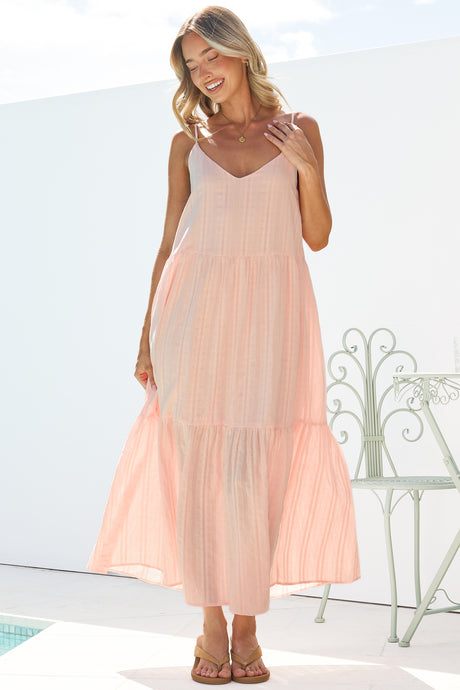 A woman twirling the skirt of the Alora Midi Dress in Pink, showing the flowy tiered skirt, textured fabric and thin adjustable straps, paired with tan sandals on a bright white terrace with a garden chair