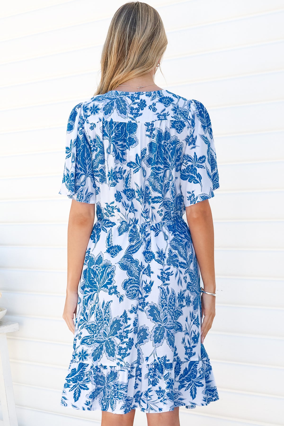 Back view of a woman wearing the Arabella Mini Dress in Alina Print, showing the flutter sleeves, gathered waist and frill hem of the blue and white floral dress, against a white wall