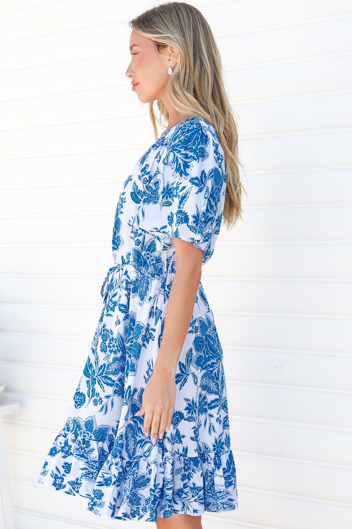 Side view of a woman wearing the Arabella Mini Dress in Alina Print, showing the flutter sleeves, tie waist and frill hem of the blue and white floral dress, against a white wall