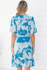 Back view of a woman wearing the Arabella Mini Dress in Colter Print Blue, showing the flutter sleeves, gathered tie waist and frill hem of the pale blue tropical leaf print dress, against a white wall