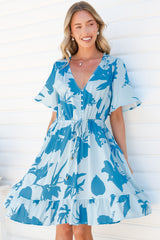 A smiling woman standing against a white wall in the Arabella Mini Dress in Colter Print Blue, showing the flowing skirt, frill hem, button front and wide flutter sleeves of the pale blue tropical leaf print dress