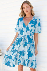 A woman holding the skirt of the Arabella Mini Dress in Colter Print Blue, a relaxed V-neck button-front dress with flutter sleeves, a drawstring tie waist and frill hem in a pale blue and bright blue tropical leaf print, against a white wall