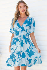 A smiling woman holding the frill hem of the Arabella Mini Dress in Colter Print Blue, a knee-length V-neck button-front dress with flutter sleeves and a drawstring tie waist in a pale blue tropical leaf print, against a white wall