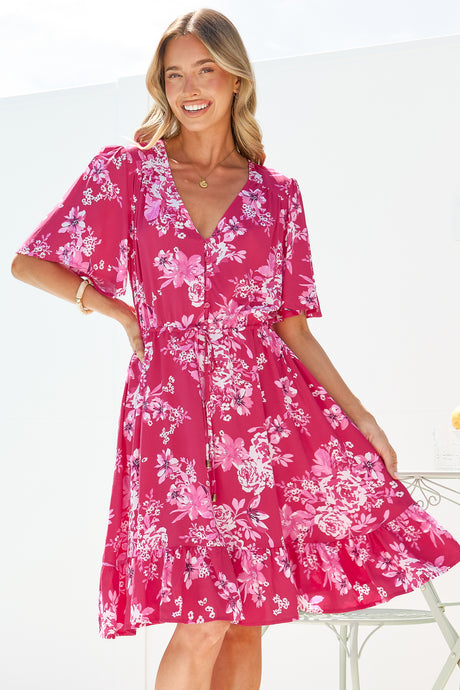 A smiling woman wearing the Arabella Mini Dress in Kimberly Print Pink, a hot pink and white floral V-neck button-front mini with flutter sleeves and a tie waist, on a bright white terrace