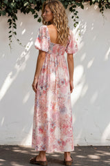 Woman wearing a floral dress standing against a white wall with greenery.