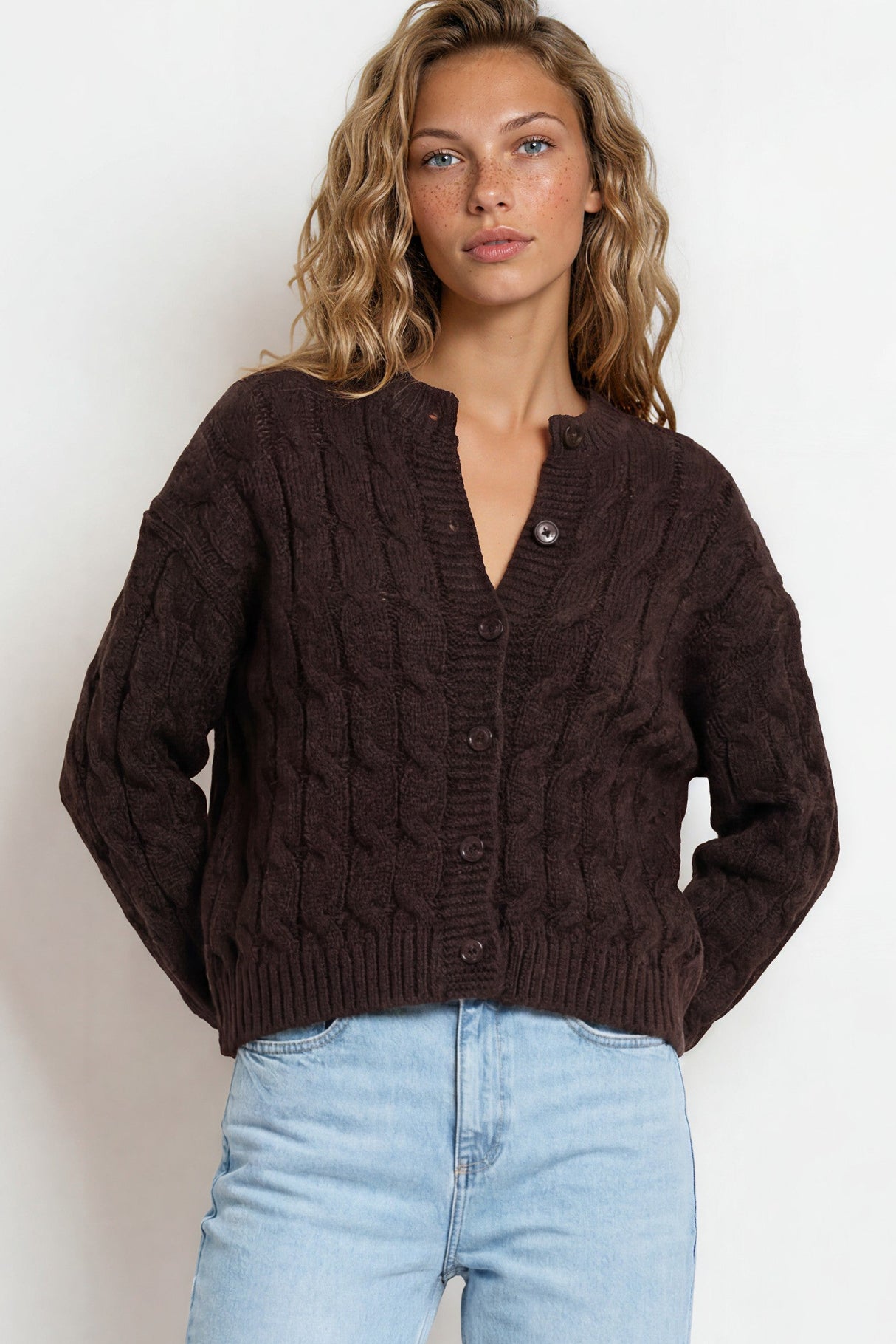 Woman wearing a brown cable knit cardigan and light blue jeans on a white background