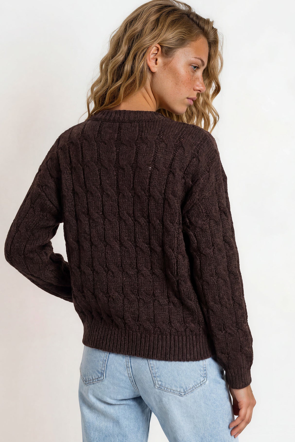 Woman wearing a brown cable knit sweater and light blue jeans on a plain background
