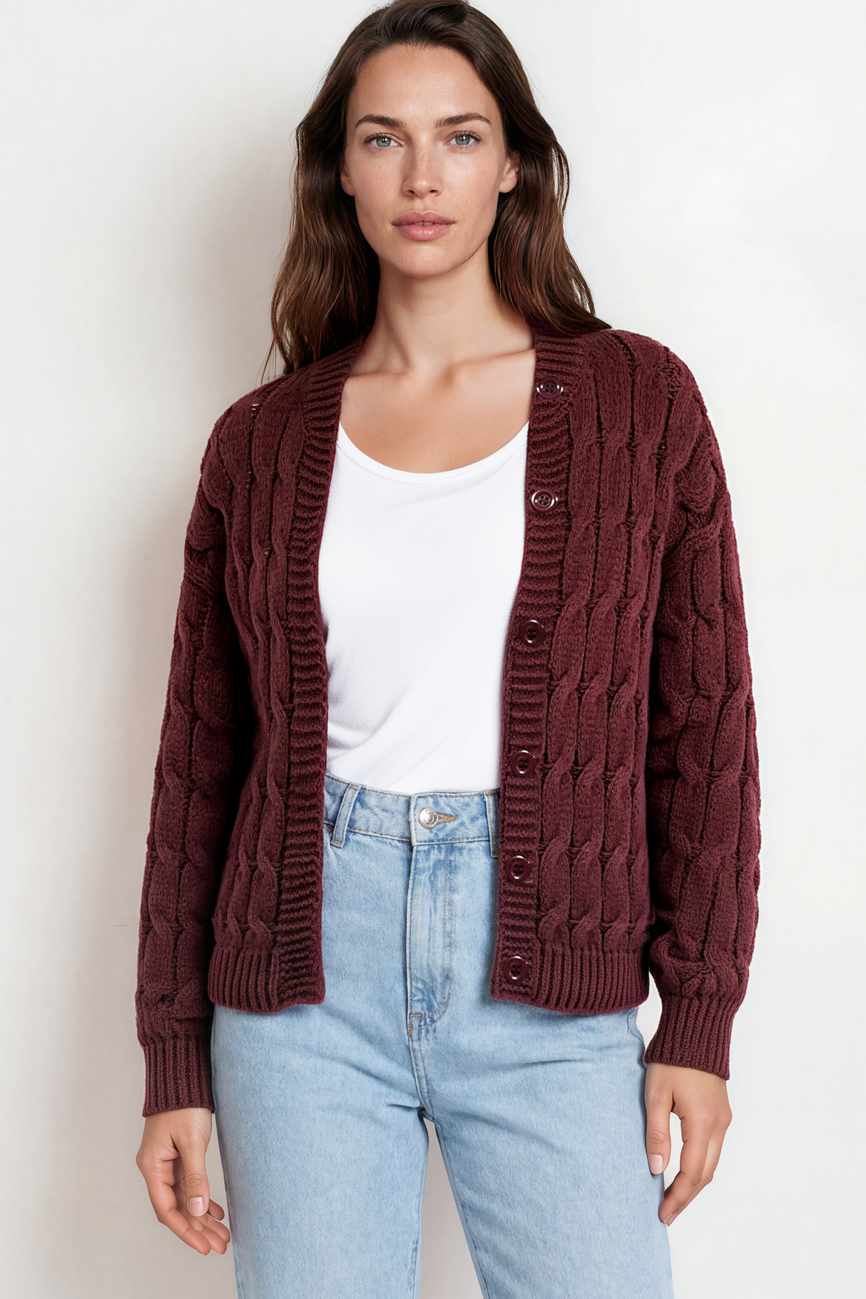 Woman wearing a maroon cable knit cardigan over a white top and light blue jeans on a plain background