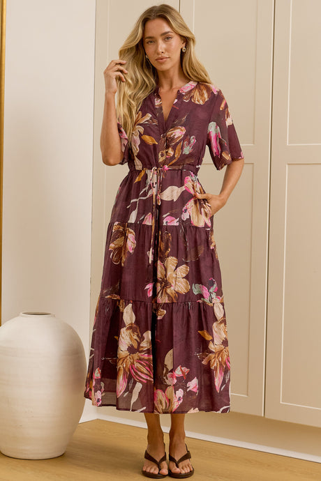 A woman stands in a bright room wearing a short-sleeve floral midi dress with a tie waist. She is positioned in front of cream-colored built-in wardrobes, with a large floor mirror on the left reflecting her side profile.