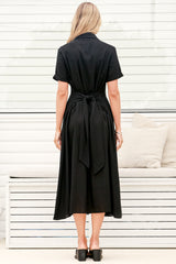 A woman wearing a black midi dress with a round neckline, short sleeves, and relaxed flowing silhouette, standing in front of a white panelled wall.