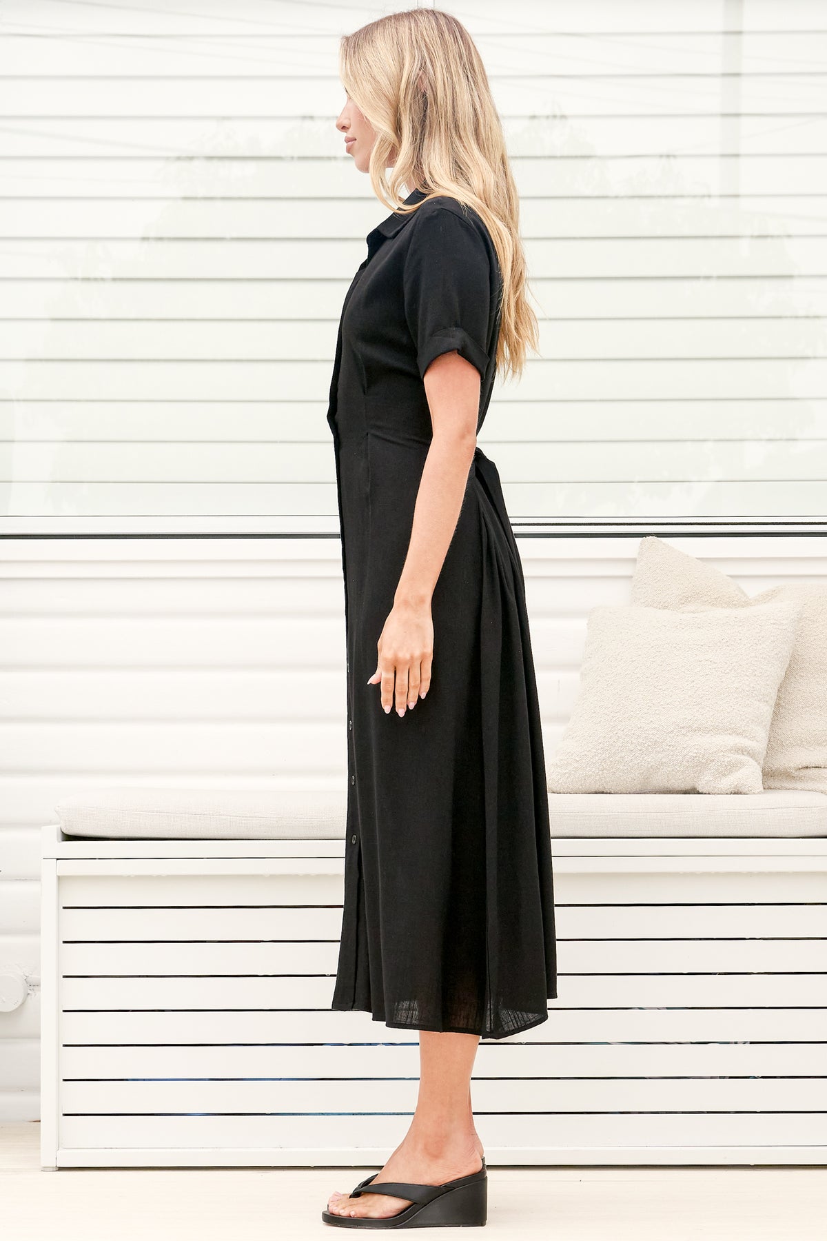 A woman wearing a black midi dress with a round neckline, short sleeves, and relaxed flowing silhouette, standing in front of a white panelled wall.