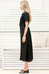 A woman wearing a black midi dress with a round neckline, short sleeves, and relaxed flowing silhouette, standing in front of a white panelled wall.