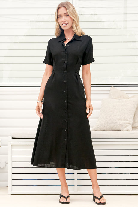 A woman wearing a black midi dress with a round neckline, short sleeves, and relaxed flowing silhouette, standing in front of a white panelled wall.