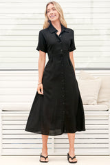 A woman wearing a black midi dress with a round neckline, short sleeves, and relaxed flowing silhouette, standing in front of a white panelled wall.