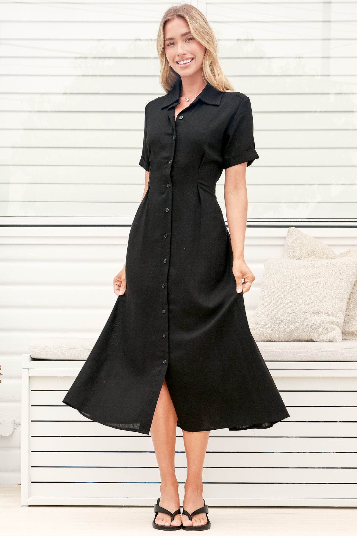 A woman wearing a black midi dress with a round neckline, short sleeves, and relaxed flowing silhouette, standing in front of a white panelled wall.