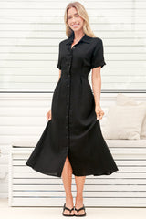 A woman wearing a black midi dress with a round neckline, short sleeves, and relaxed flowing silhouette, standing in front of a white panelled wall.