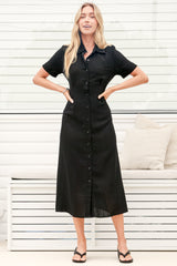 A woman wearing a black midi dress with a round neckline, short sleeves, and relaxed flowing silhouette, standing in front of a white panelled wall.