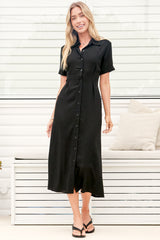 A woman wearing a black midi dress with a round neckline, short sleeves, and relaxed flowing silhouette, standing in front of a white panelled wall.