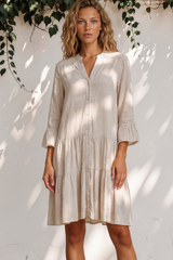 Woman wearing a light-colored dress standing against a white wall with greenery.
