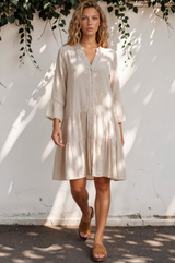 Woman wearing a beige dress standing against a white wall with greenery.