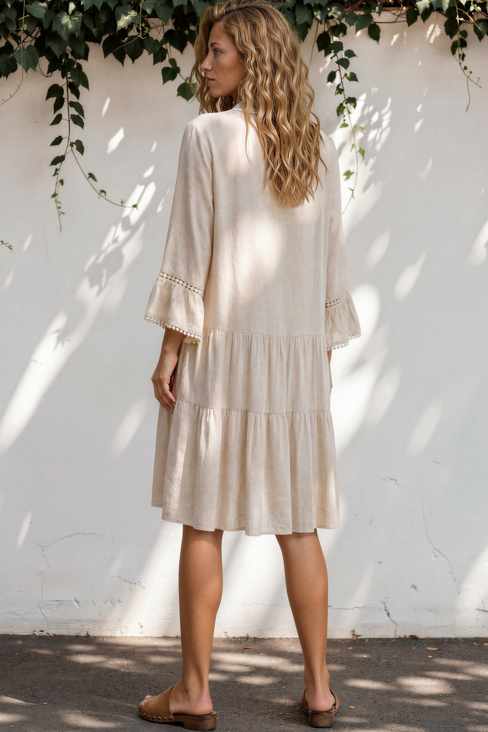 Woman wearing a beige dress standing against a white wall with greenery.