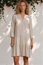 Woman wearing a beige dress standing against a white wall with greenery.