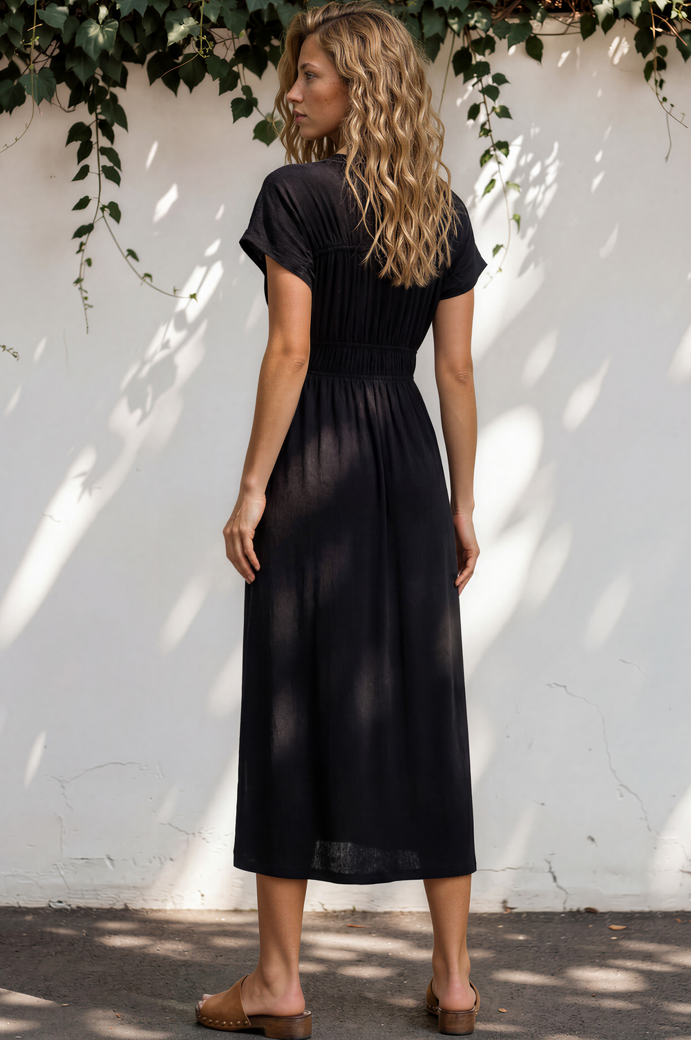 Woman in a black dress standing against a white wall with greenery.