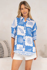 A woman wearing the Blaze Shirt in Atrina Print, a collared short-sleeve button-down with a blue and white seashell print, styled with white shorts in front of a cream sofa with neutral cushions